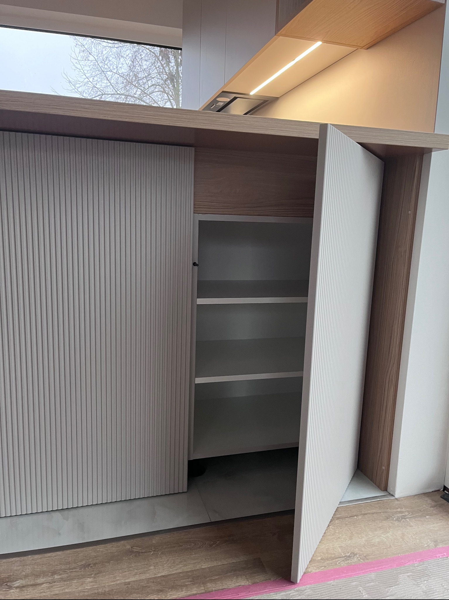 Nowoczesna szafka kuchenna z ryflowanym frontem w odcieniach bieli i drewna, częściowo otwarta, ukazująca półki. Minimalistyczny design, jasne wnętrze.