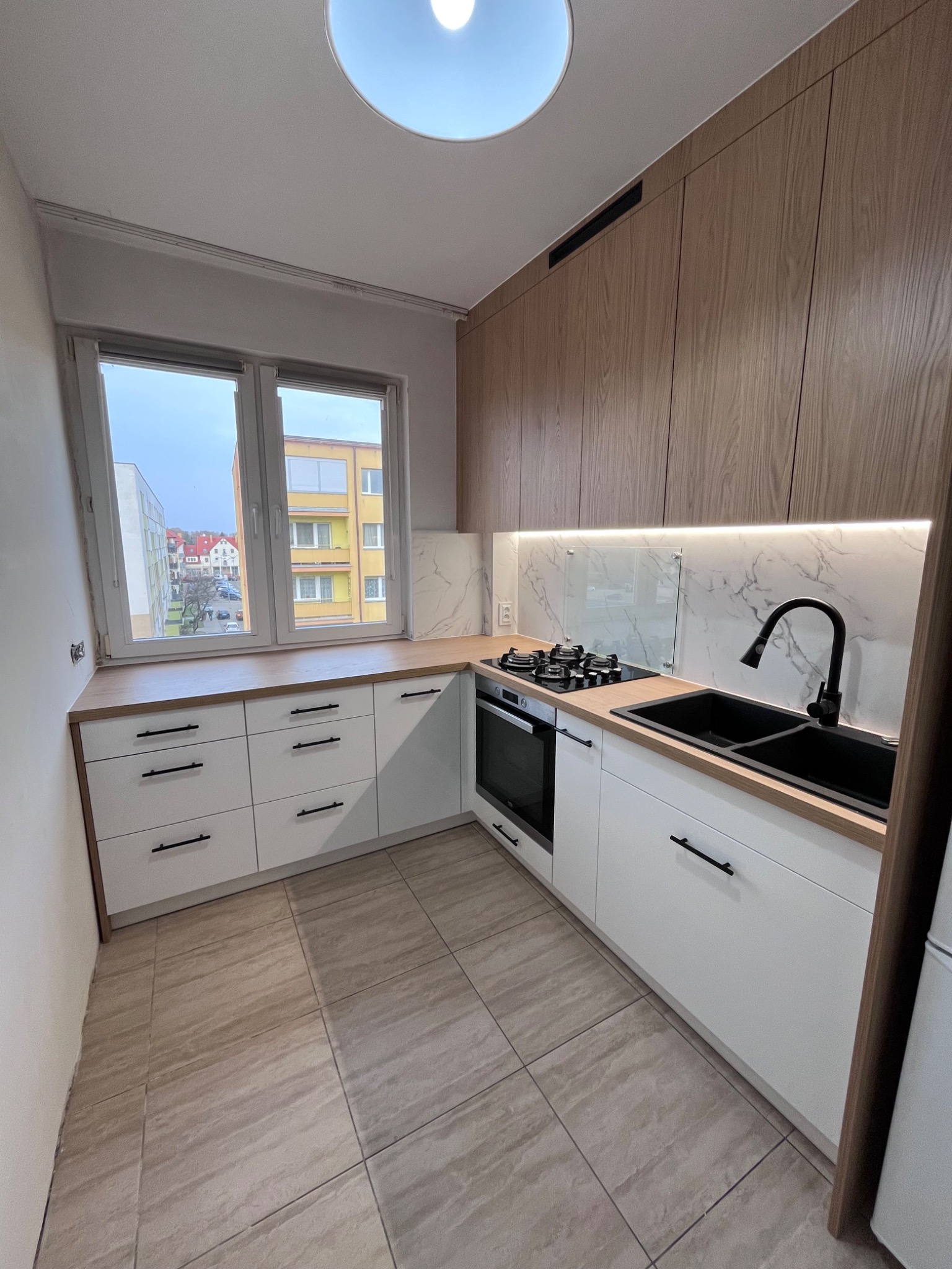 Nowoczesna kuchnia z białymi szafkami i drewnianymi frontami górnymi. Czarny zlew i bateria, marmurowy backsplash, płyta grzewcza. Widok z okna na budynki.