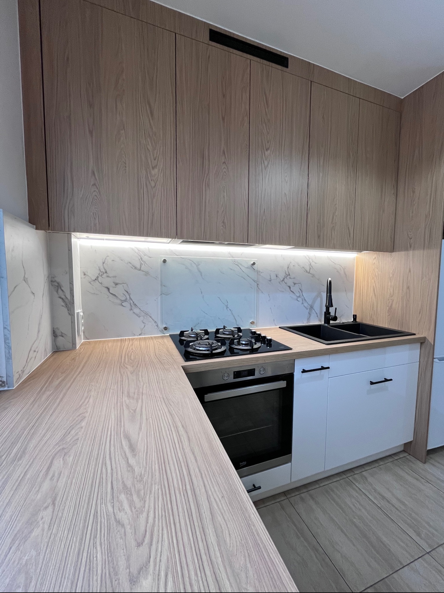 Jasna kuchnia z drewnianymi szafkami górnymi i białymi dolnymi. Blat i ściana imitują marmur. Czarny zlew i bateria. Nowoczesny design z oświetleniem LED.