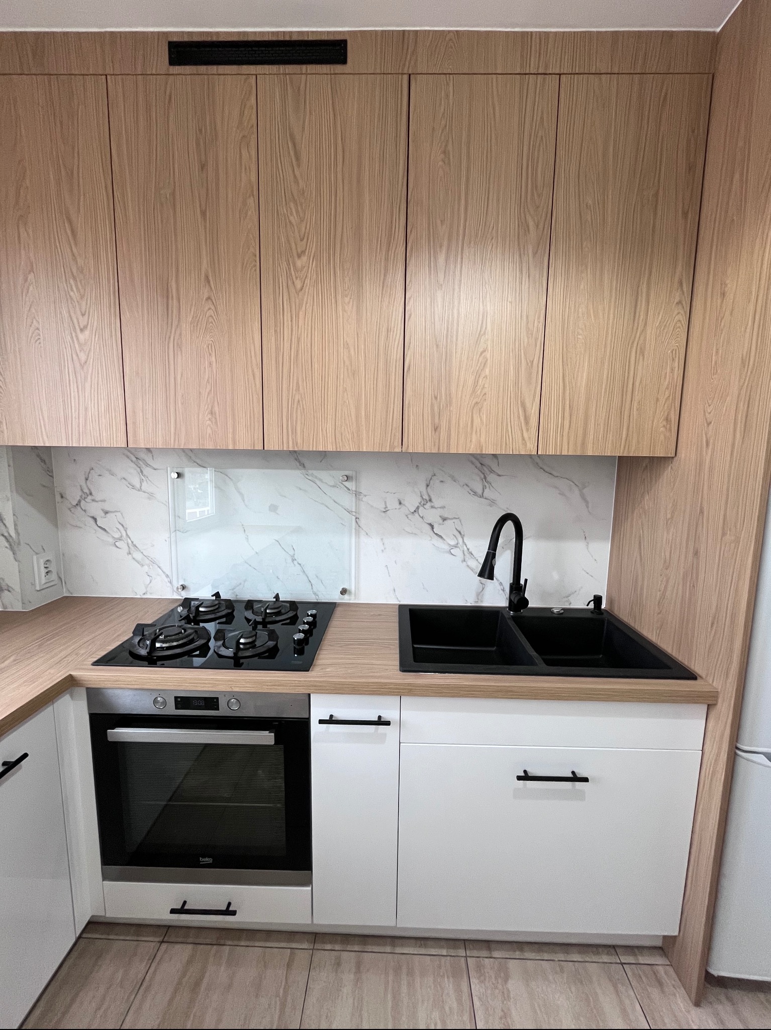 Jasna kuchnia z drewnianymi szafkami górnymi i białymi dolnymi. Czarny zlew i płyta gazowa. Marmurowy backsplash za płytą. Minimalistyczne uchwyty.