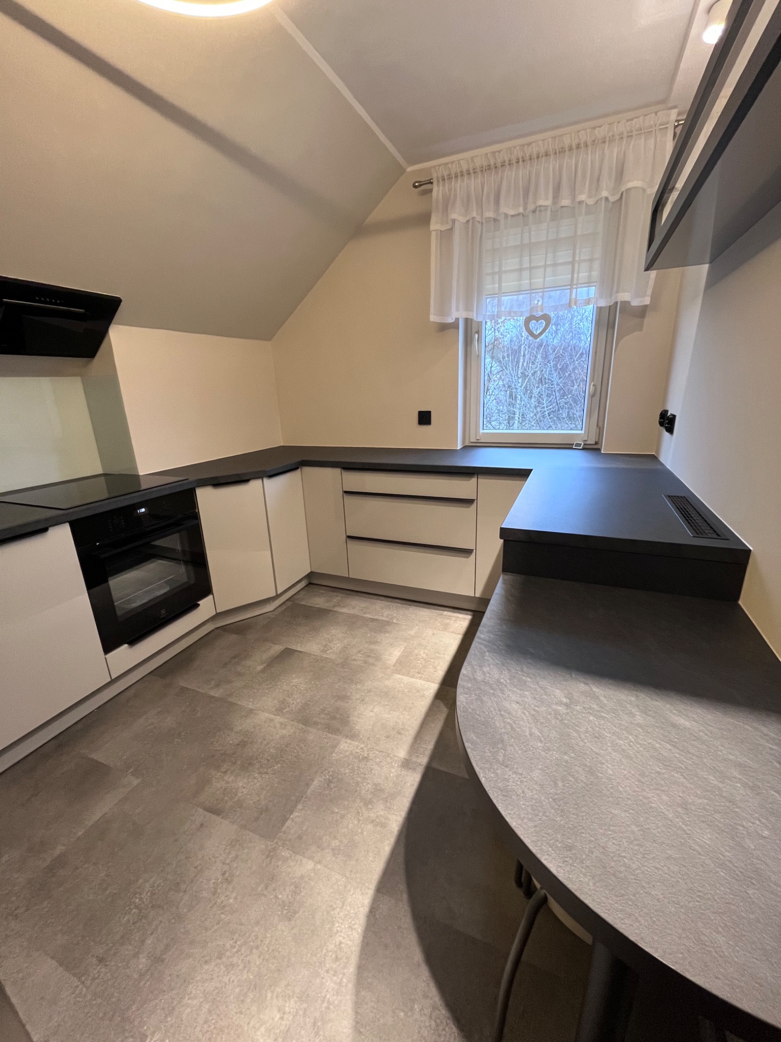 Jasna kuchnia na poddaszu z szarym blatem i białymi frontami. Okno z firanką i serduszkiem. Płyta indukcyjna i piekarnik w zabudowie. Nowoczesny design.