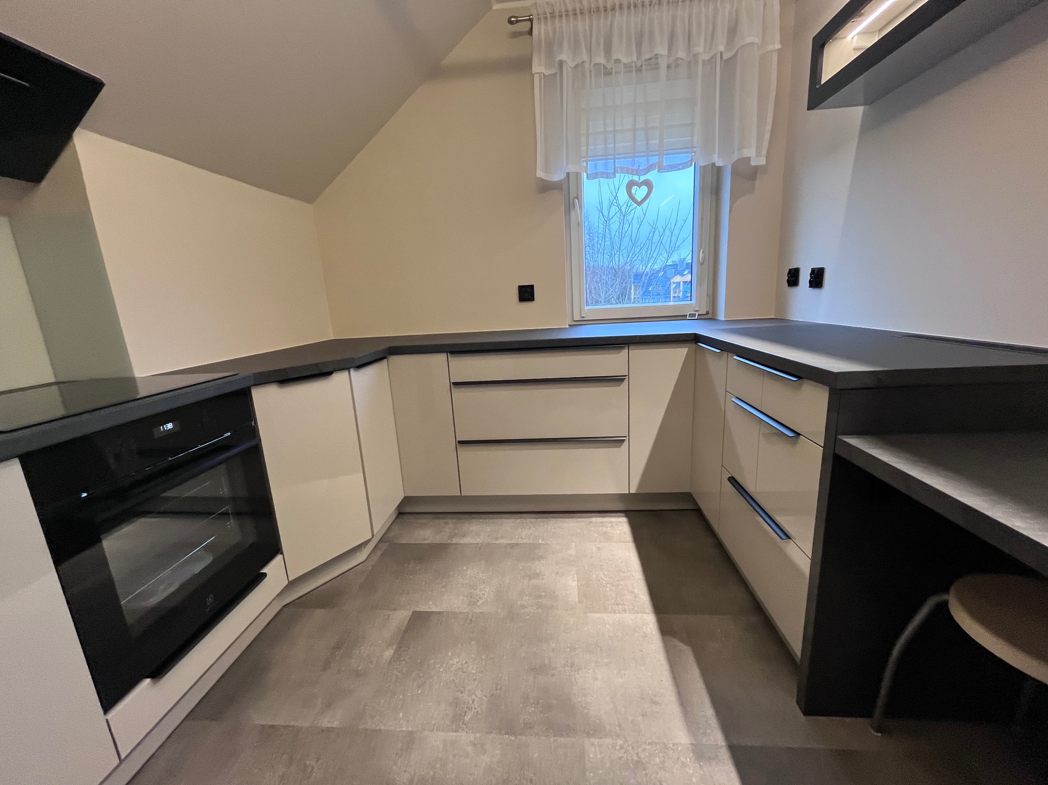 Jasna kuchnia z ciemnym blatem w Gryfinie. Zabudowa pod skosem, okno z serduszkiem. Nowoczesny design, minimalistyczne uchwyty, piekarnik w zabudowie.