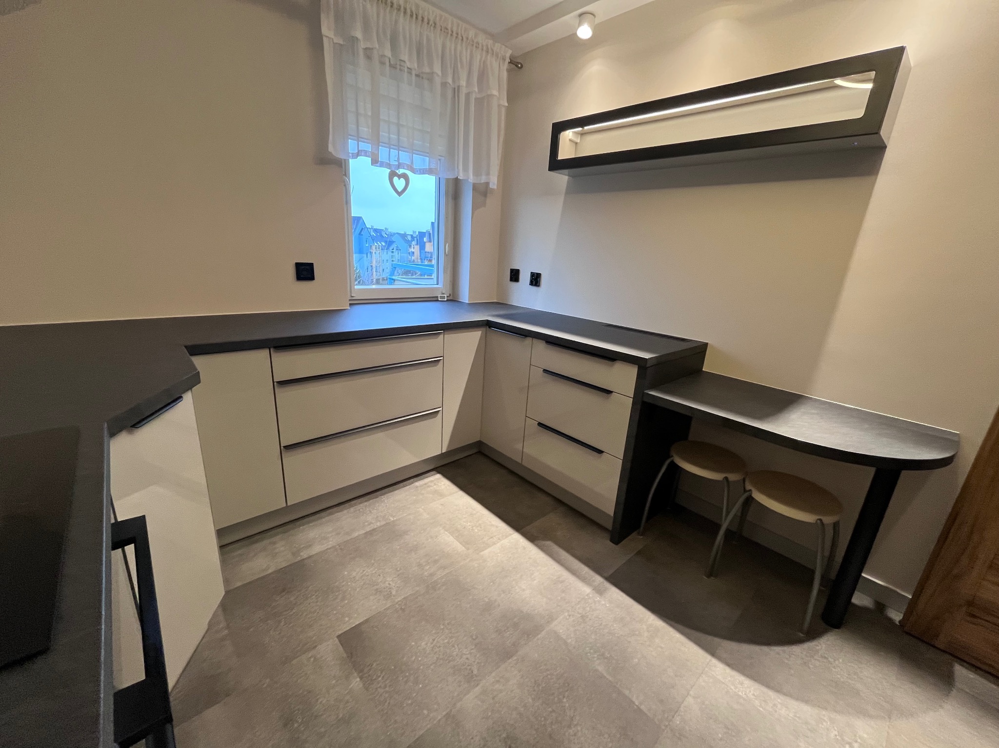 Jasna kuchnia z szarym blatem, minimalistyczne fronty szafek z poziomymi uchwytami, okno z firanką i serduszkiem, dodatkowe oświetlenie nad blatem.