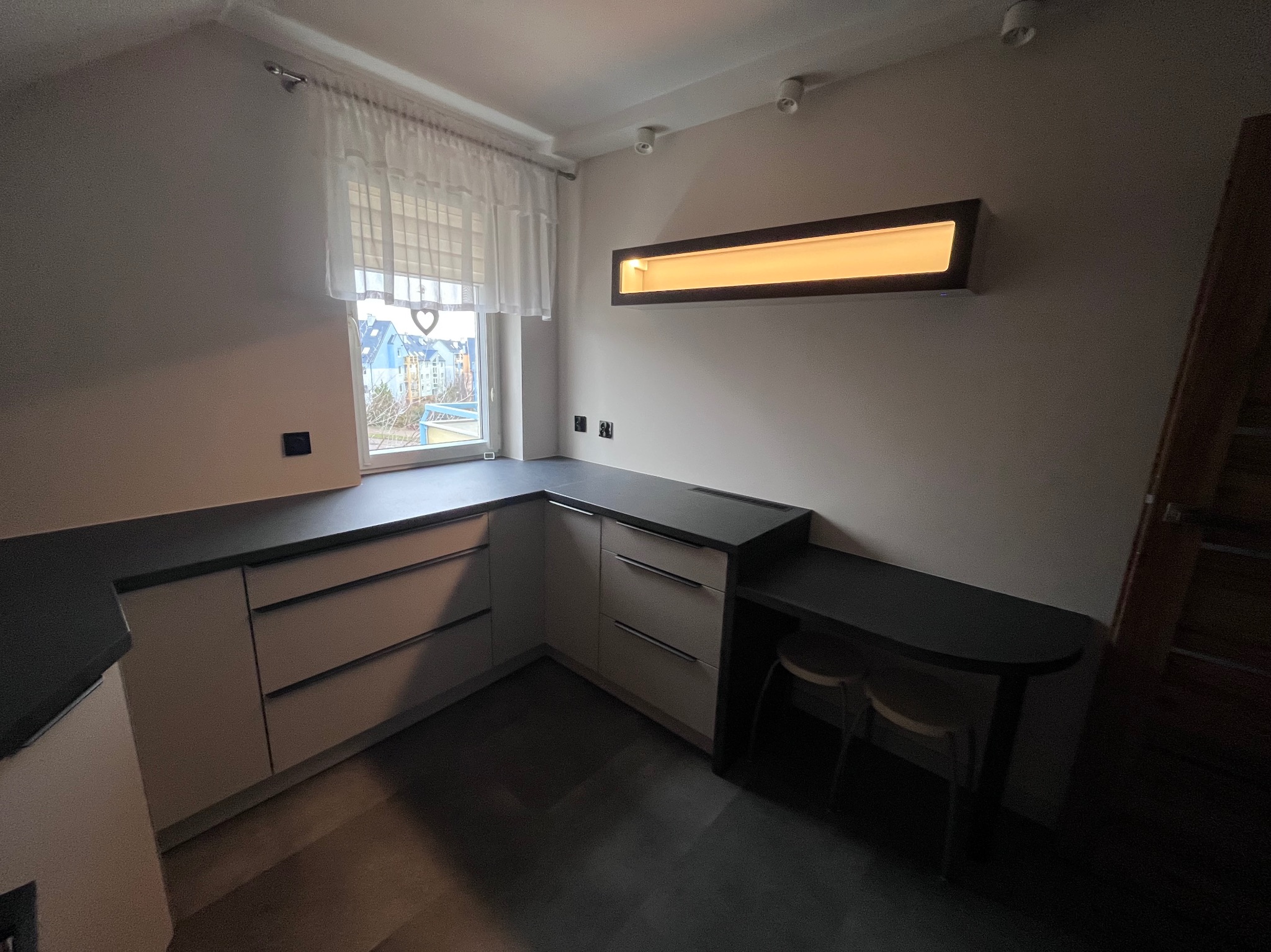 Nowoczesna kuchnia na poddaszu z szarymi frontami, blatem i podłogą. Okno z białą firanką i podświetlana wnęka nad blatem. Dwa krzesła przy blacie.