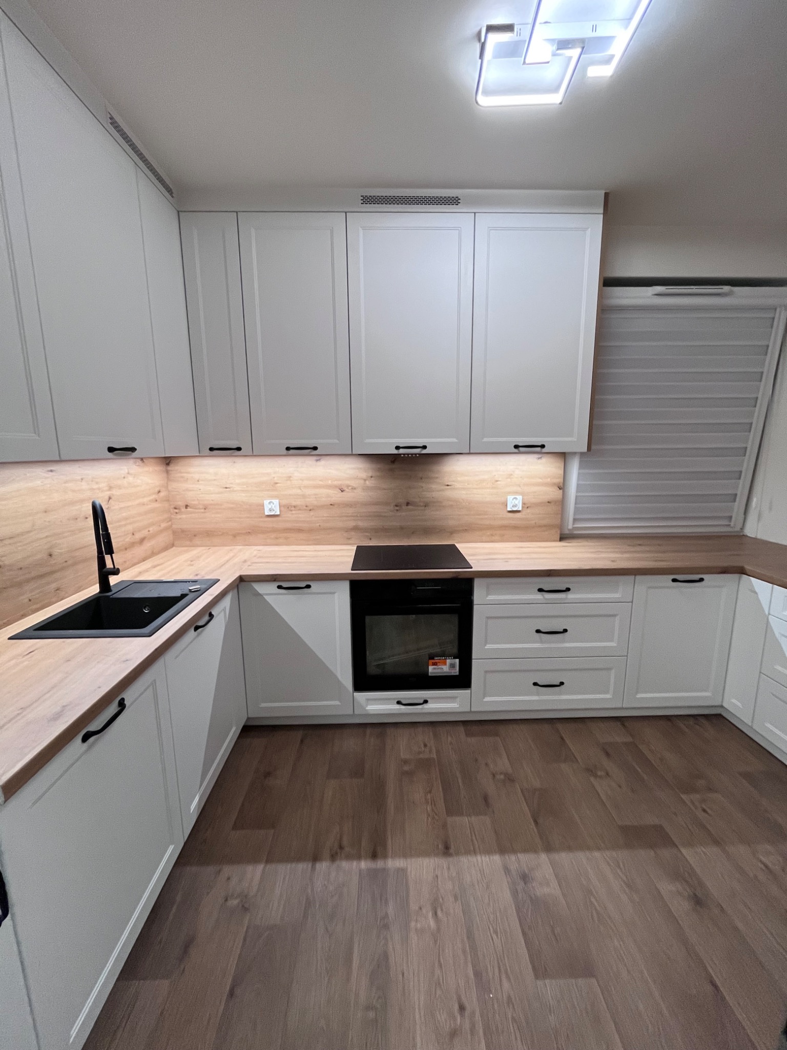 Jasna kuchnia na wymiar z drewnianym blatem i czarną armaturą. Górne szafki z frezowanymi frontami. Widoczna płyta indukcyjna i okno z żaluzjami.