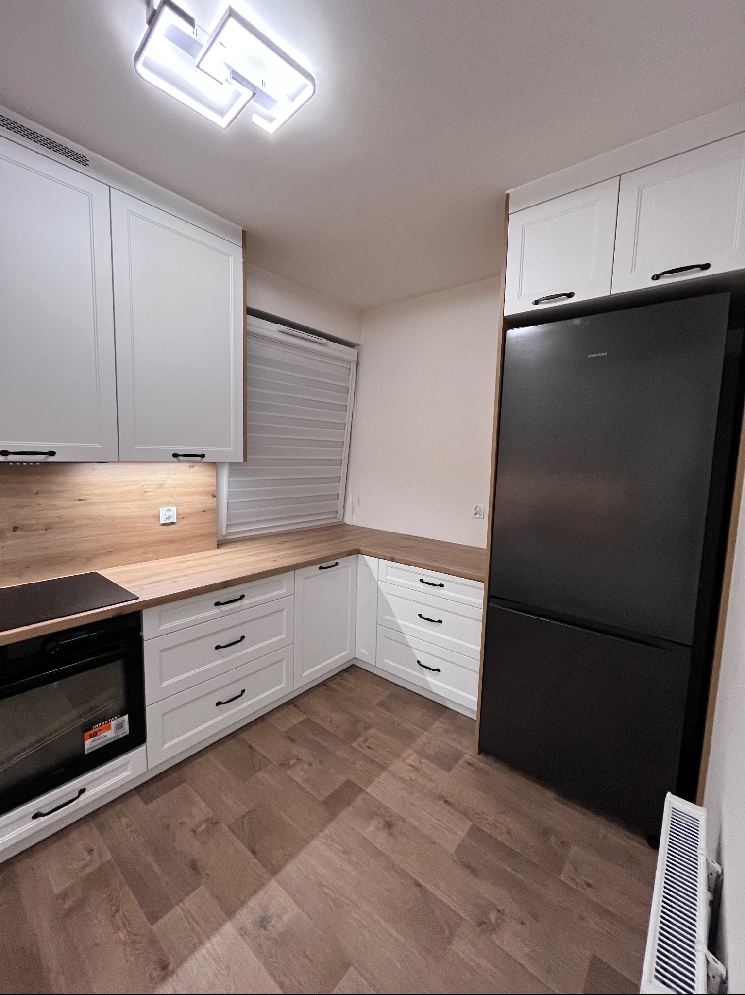 Biała kuchnia z drewnianym blatem i czarnymi uchwytami, oświetlona lampą sufitową. W tle żaluzje okienne i czarna lodówka Samsung.