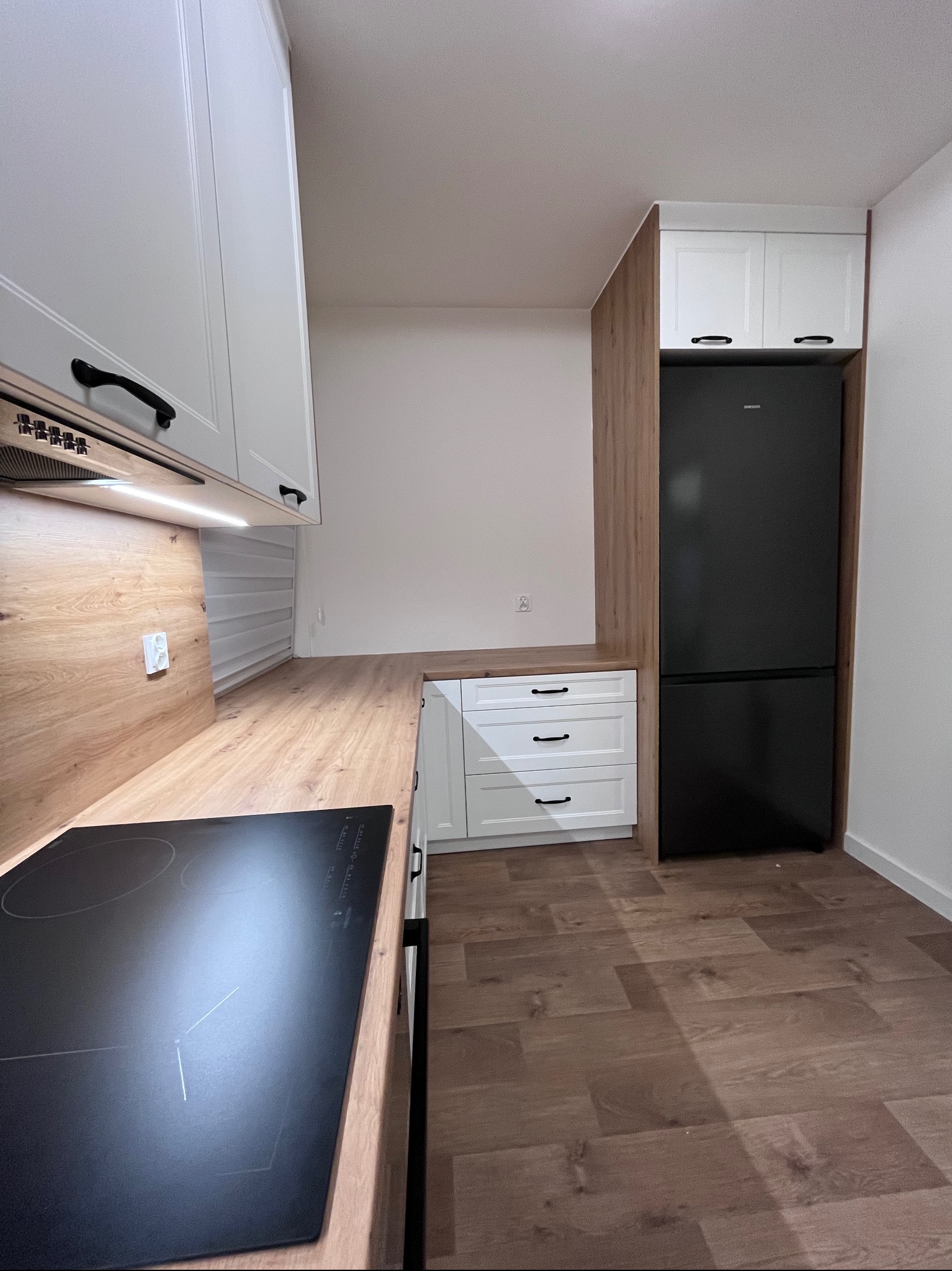 Nowoczesna kuchnia z białymi szafkami i drewnianym blatem. Czarna płyta indukcyjna i lodówka w zabudowie. Minimalistyczny design z akcentami drewna.