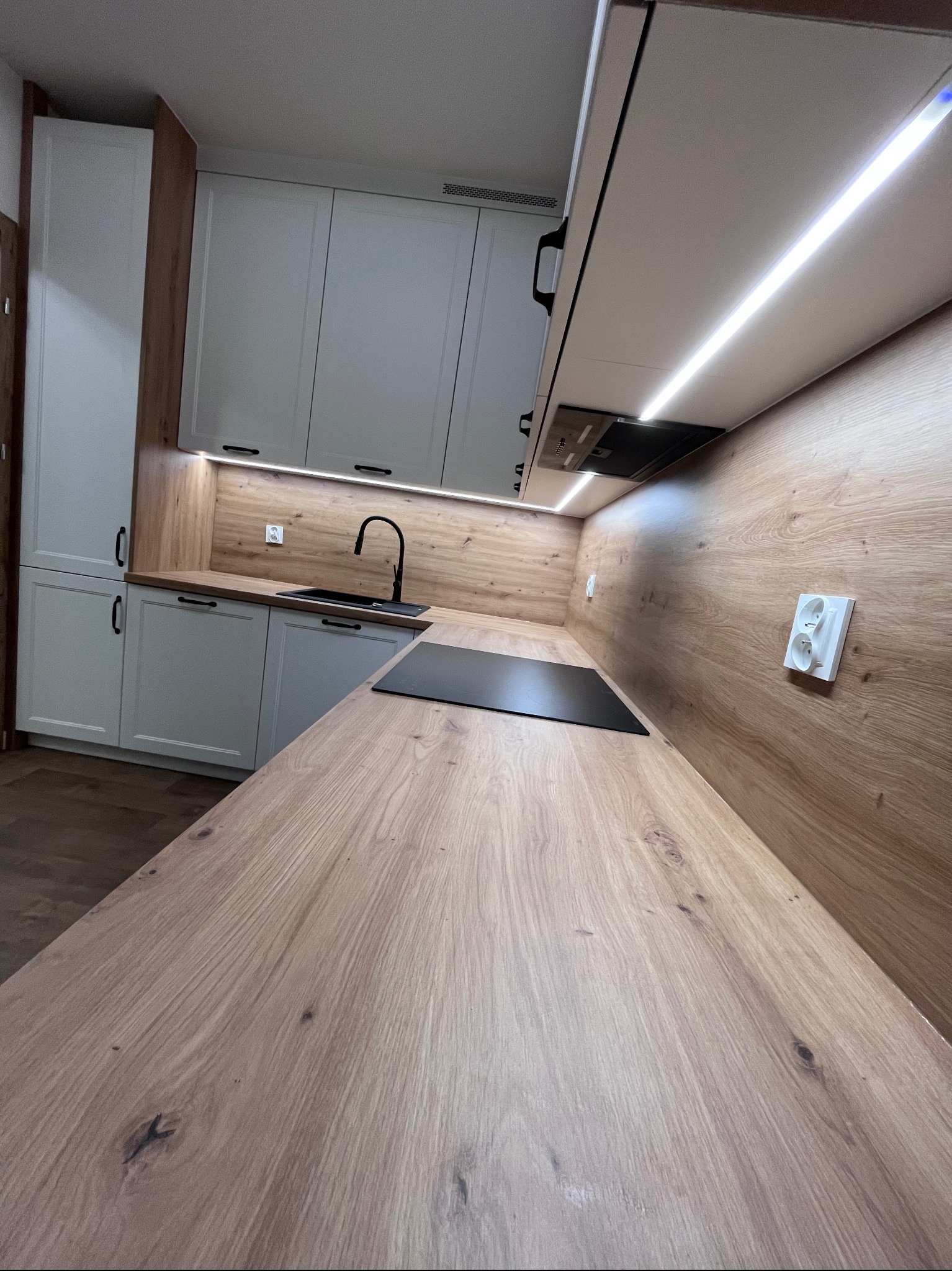 Nowoczesna kuchnia z białymi szafkami i drewnianym blatem. Uwagę przykuwa oświetlenie LED pod szafkami wiszącymi i czarna armatura.