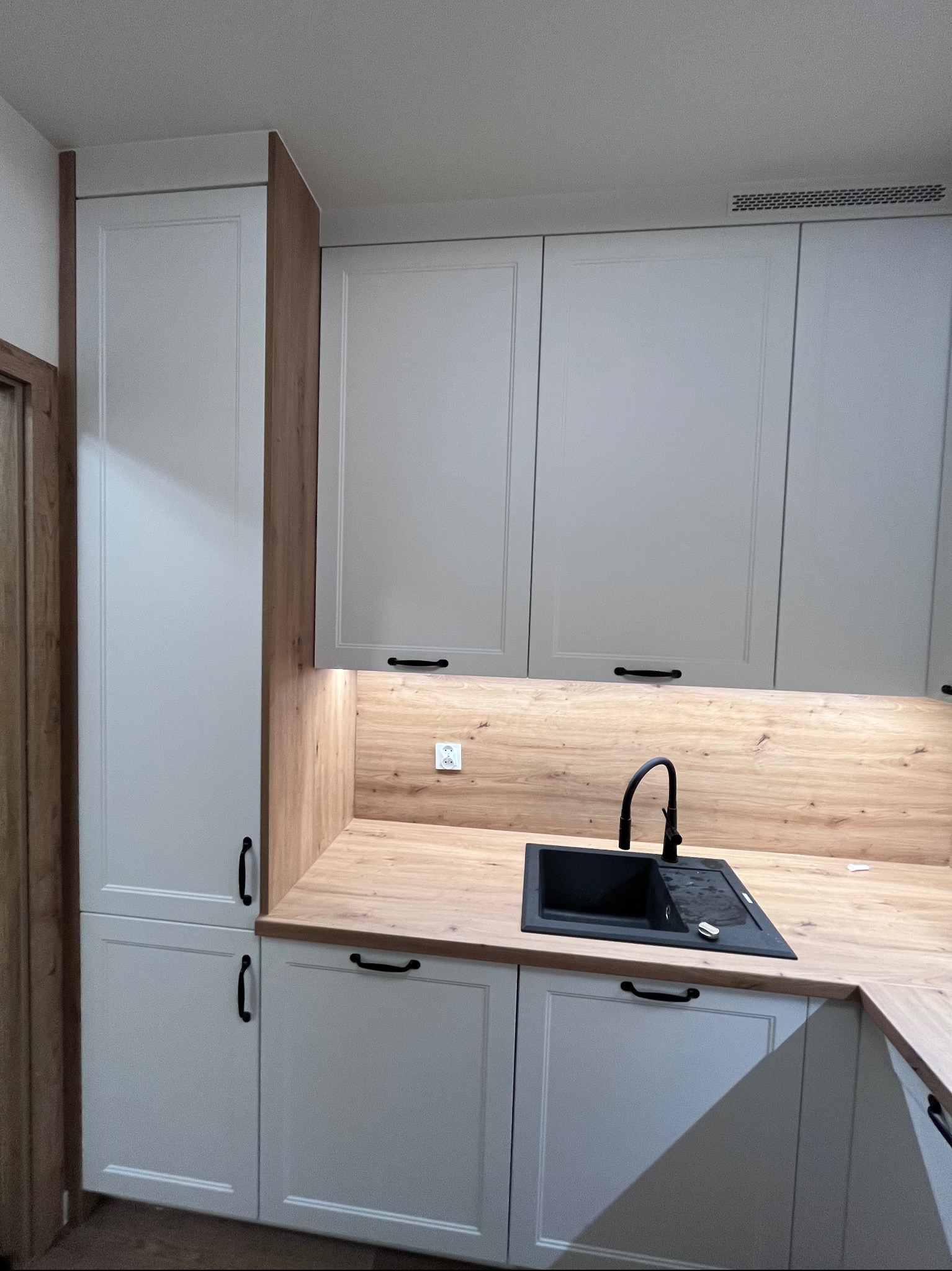 Jasna kuchnia z drewnianym blatem i czarnym zlewem. Fronty szafek w stylu klasycznym, z delikatnym frezem. Oświetlenie LED pod szafkami górnymi.