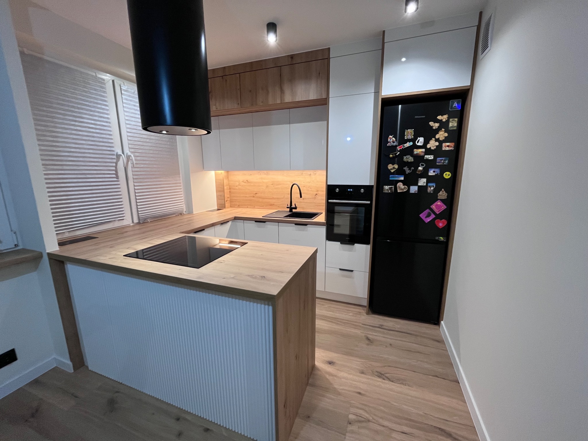Nowoczesna kuchnia z wyspą i czarnym okapem cylindrycznym. Białe fronty szafek kontrastują z drewnianym blatem i panelami. Czarna lodówka z magnesami.