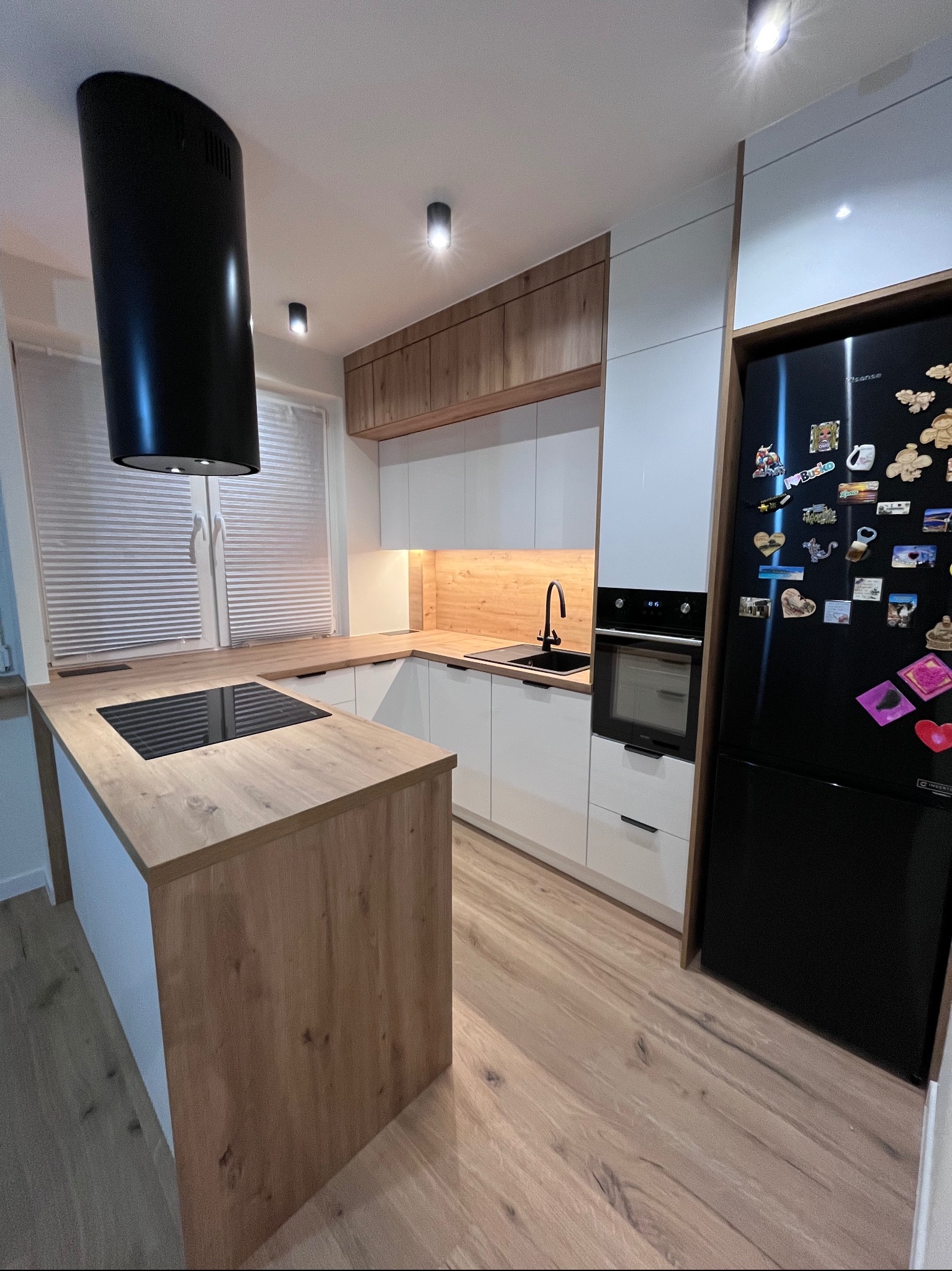 Nowoczesna kuchnia z wyspą: białe szafki, drewniane blaty, czarny okap cylindryczny i lodówka z magnesami. Minimalistyczny design, ciepłe oświetlenie.