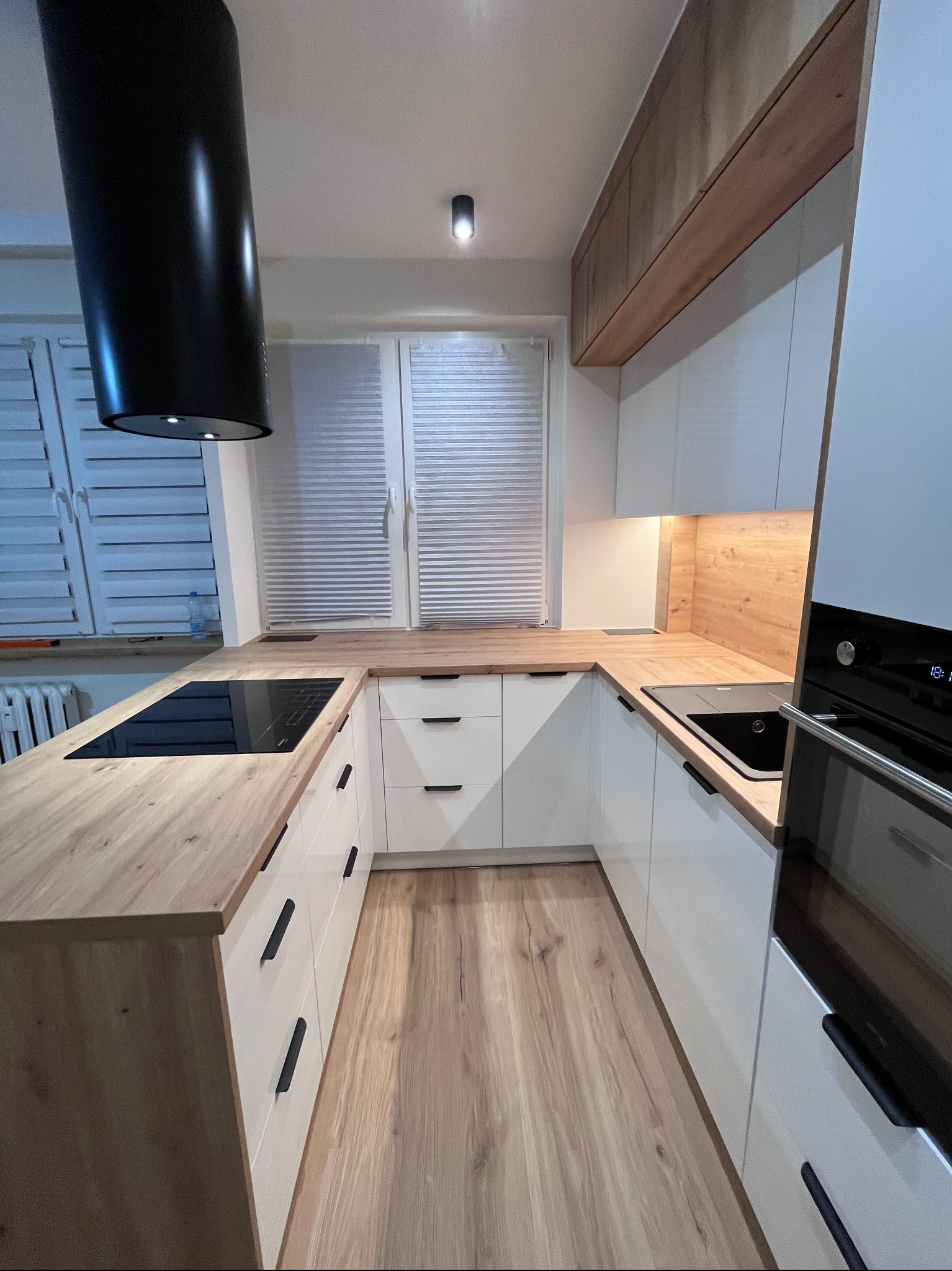 Nowoczesna kuchnia na wymiar z białymi frontami i drewnianym blatem. Czarny okap cylindryczny i minimalistyczne uchwyty. Ujęcie z perspektywy podłogi.