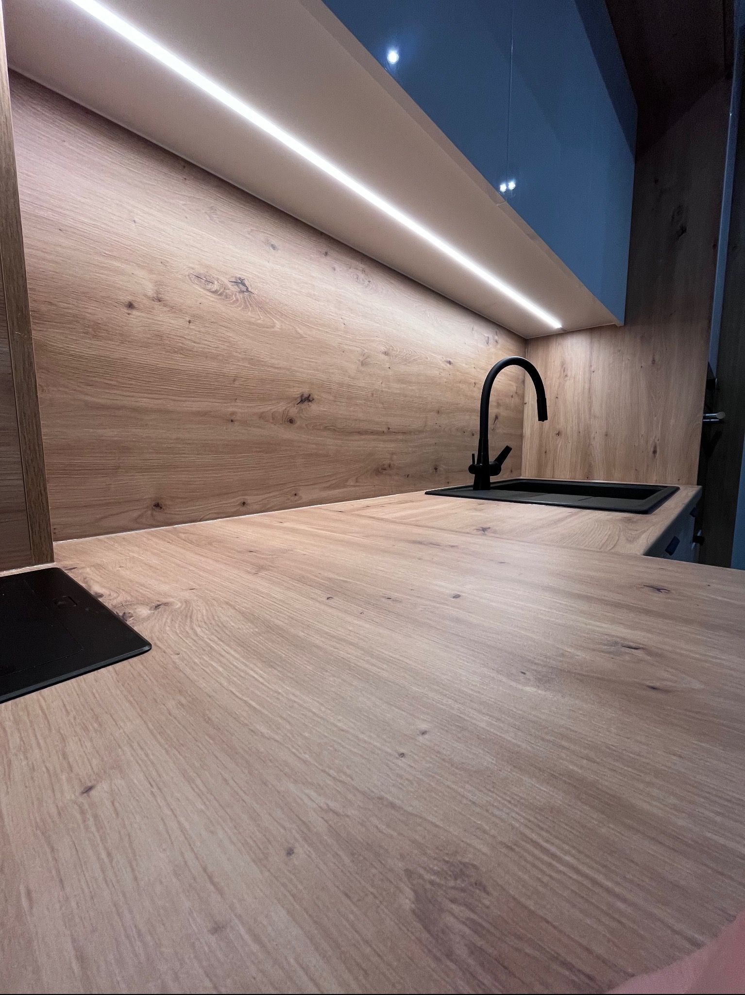 Nowoczesna kuchnia z drewnianym blatem i oświetleniem LED. Czarna bateria i zlew dodają elegancji. Widoczny fragment płyty indukcyjnej. Ujęcie z niskiej perspektywy.
