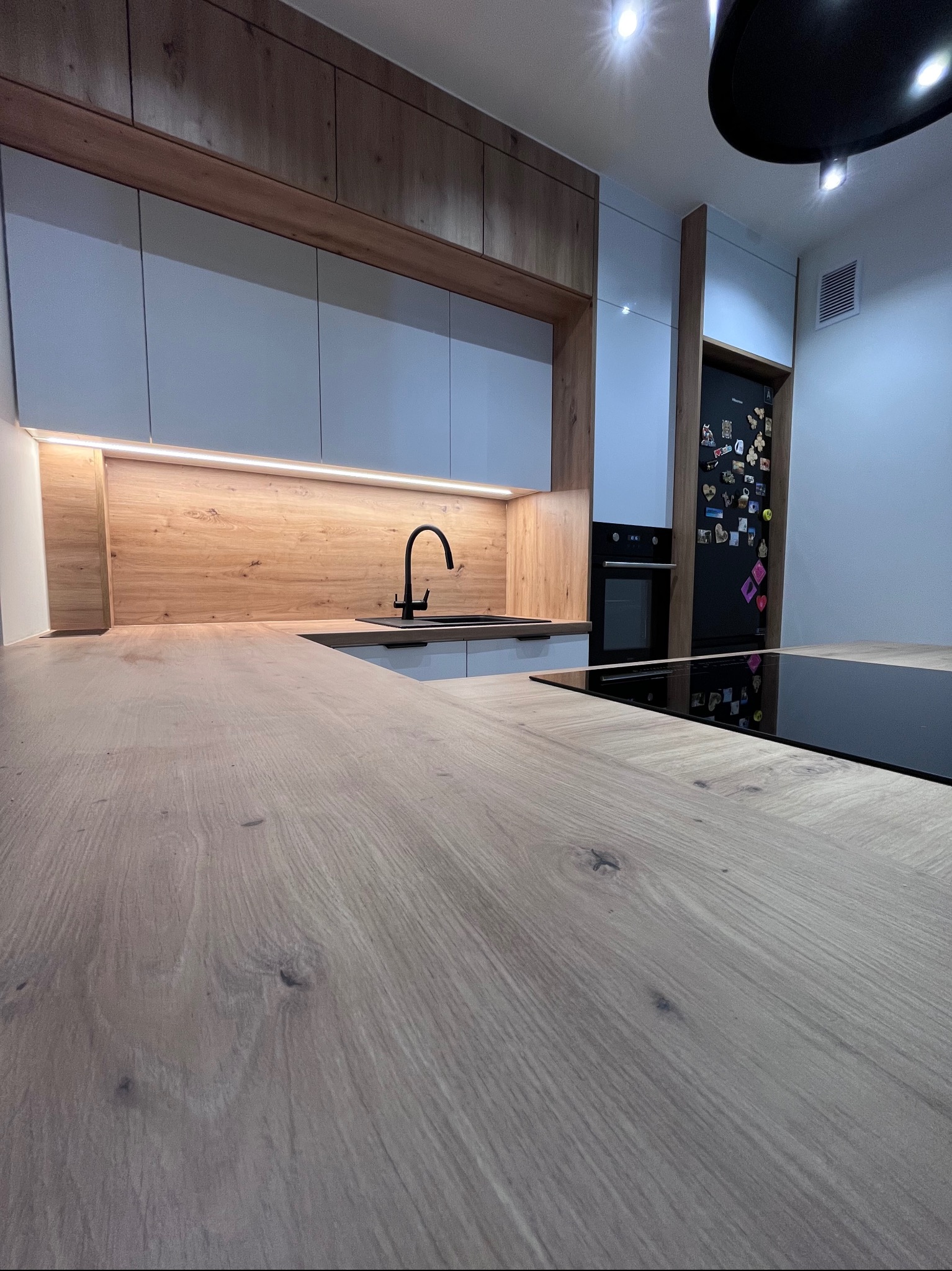 Nowoczesna kuchnia z drewnianym blatem i białymi szafkami. Czarny kran i płyta indukcyjna. Lodówka z magnesami. Oświetlenie LED pod szafkami.