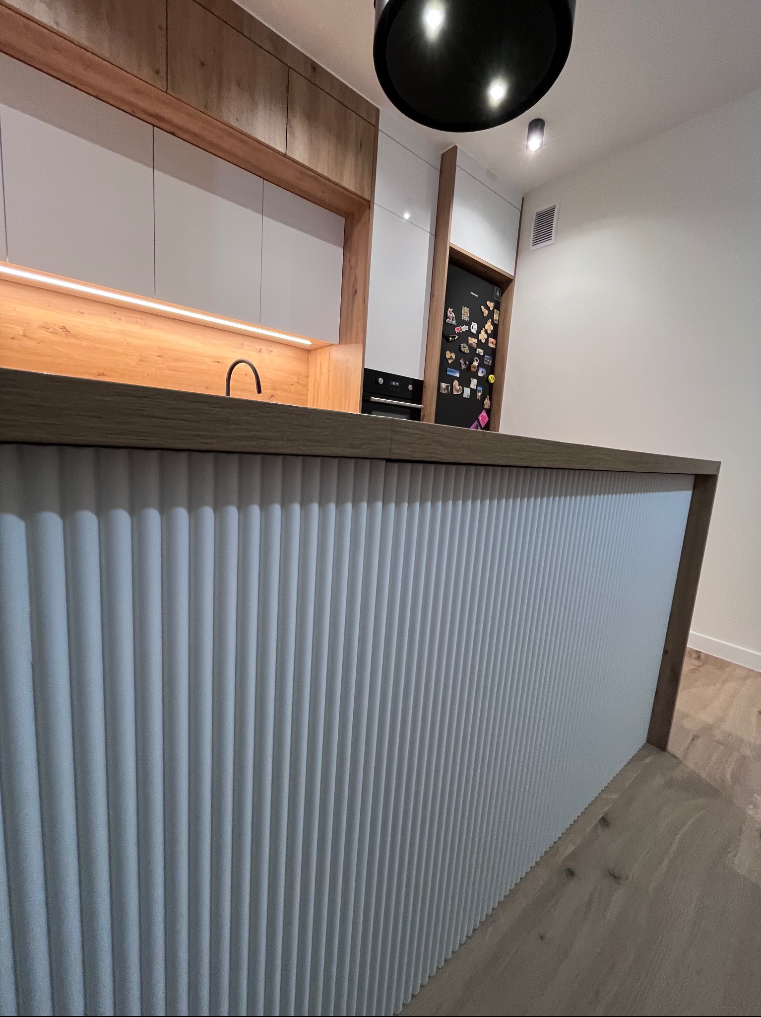 Nowoczesna kuchnia z wyspą: białe, ryflowane fronty, drewniane akcenty, czarna armatura i oświetlenie. Minimalistyczny design, ciepłe światło LED pod szafkami.