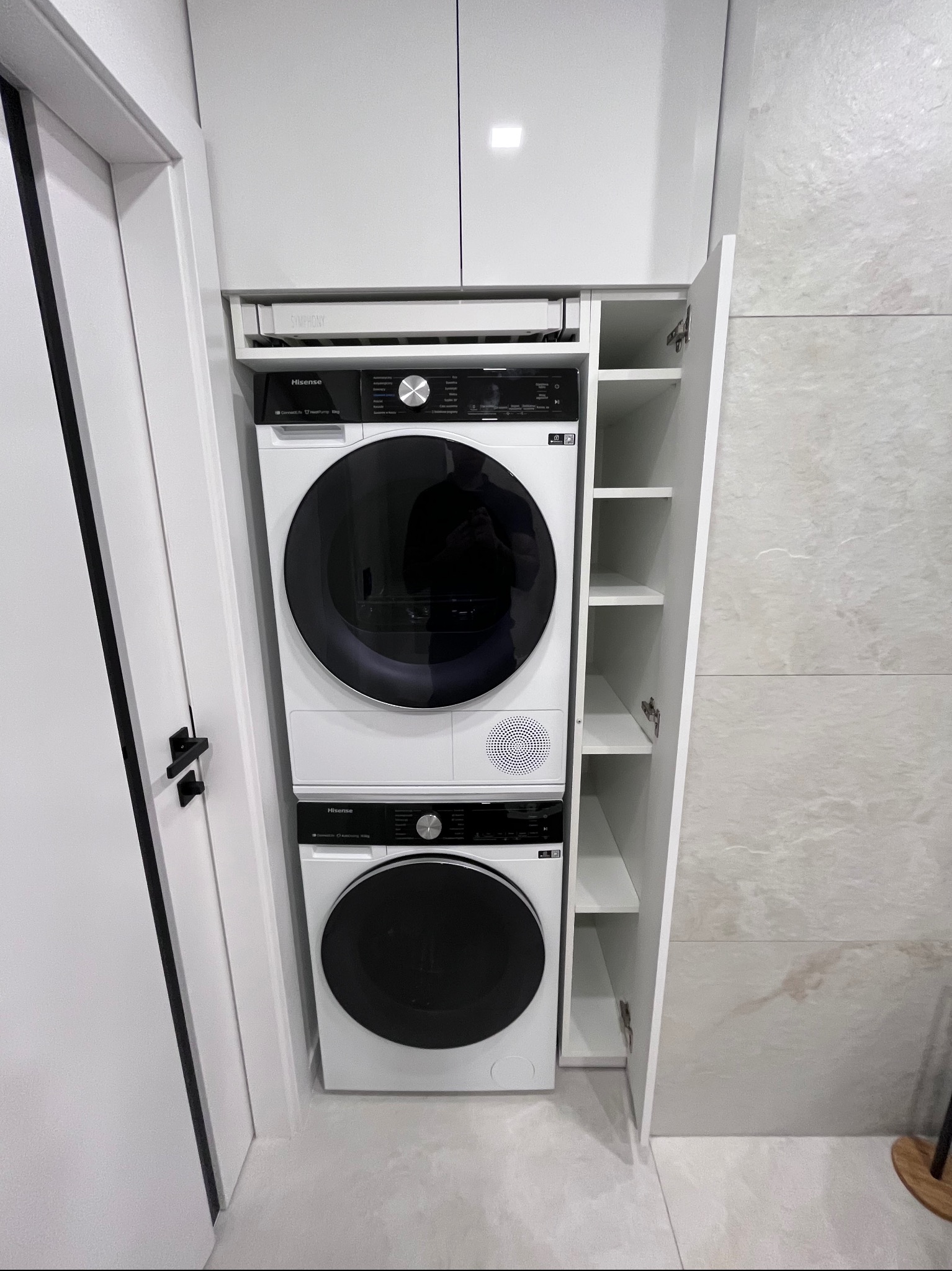 Biała zabudowa z pralką i suszarką Hisense w kolumnie, obok szafka z półkami. Minimalistyczny design, jasne wnętrze, płytki imitujące kamień na ścianie.