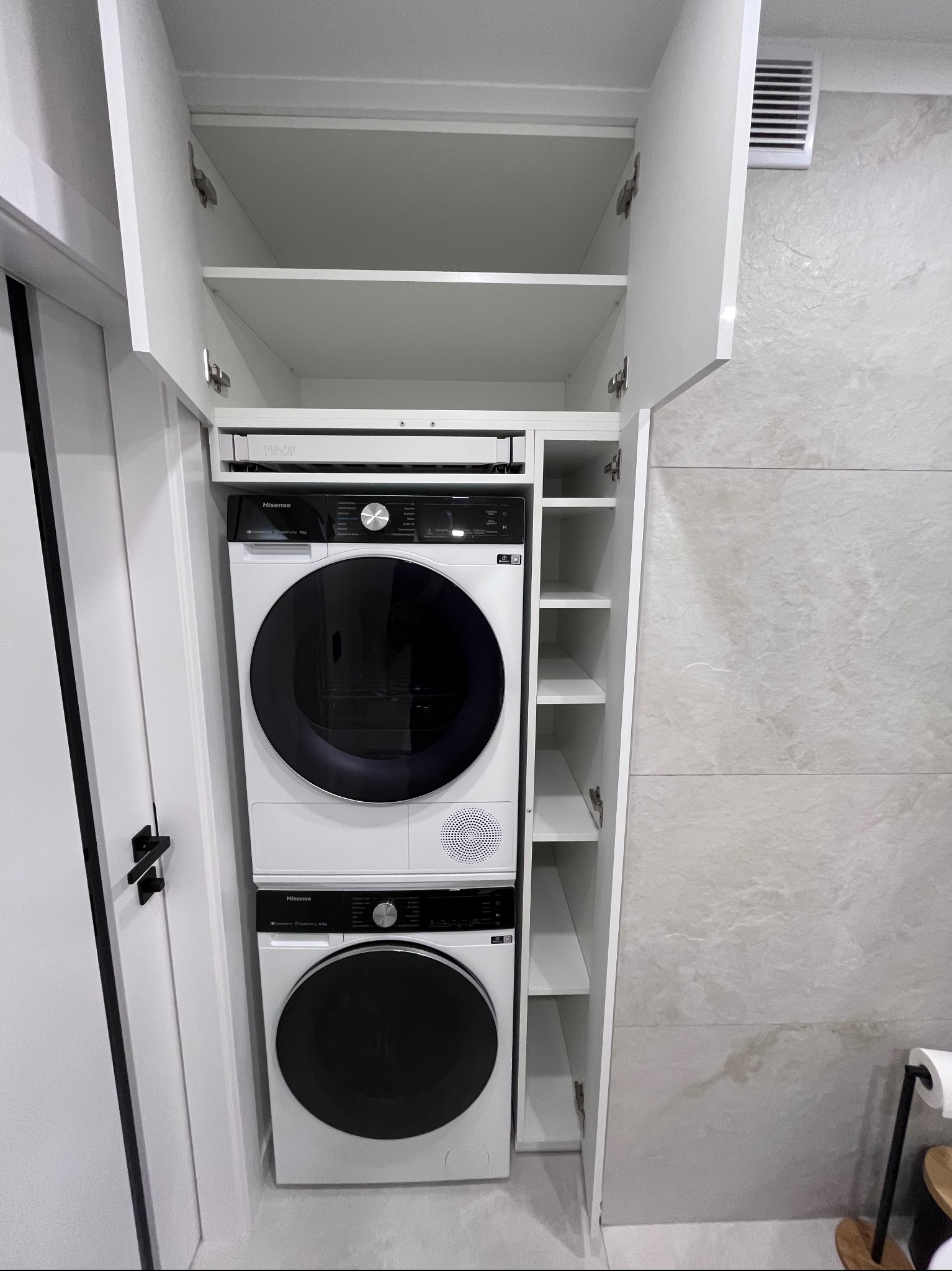 Biała zabudowa meblowa z pralką i suszarką w łazience. Otwarta szafka z półkami po prawej stronie. Minimalistyczny design, jasne płytki na ścianie.