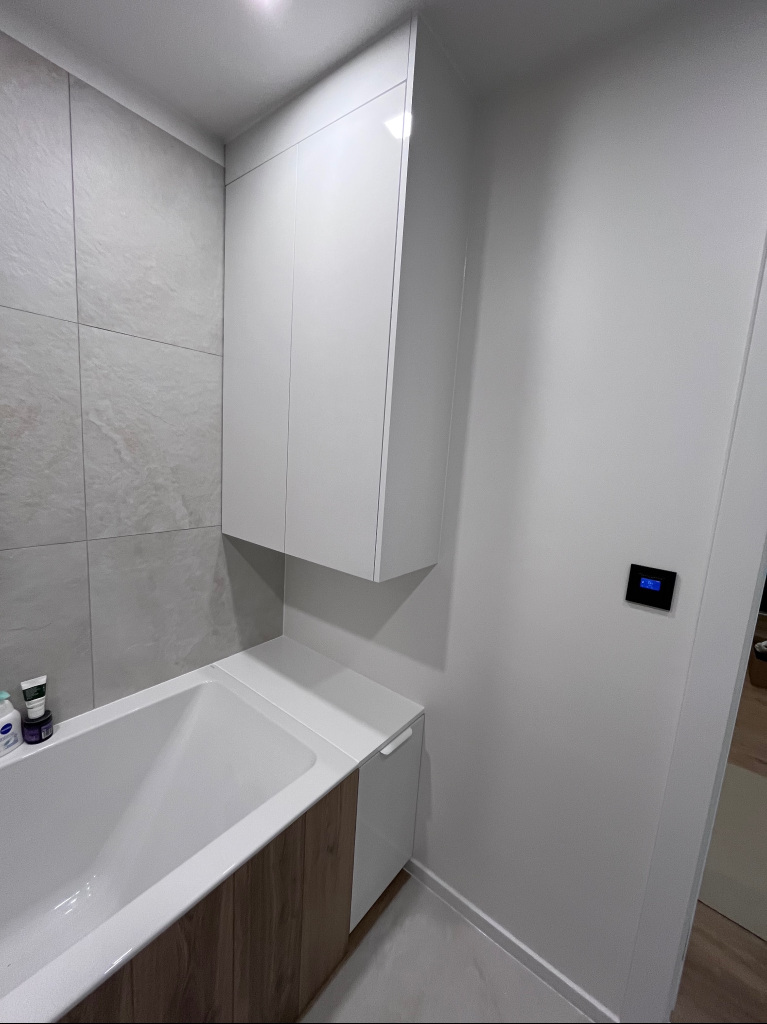 Nowoczesna łazienka z zabudową wanny z drewnopodobnym frontem i białymi szafkami wiszącymi. Minimalistyczny design, szare płytki na ścianie, czarny panel sterowania.