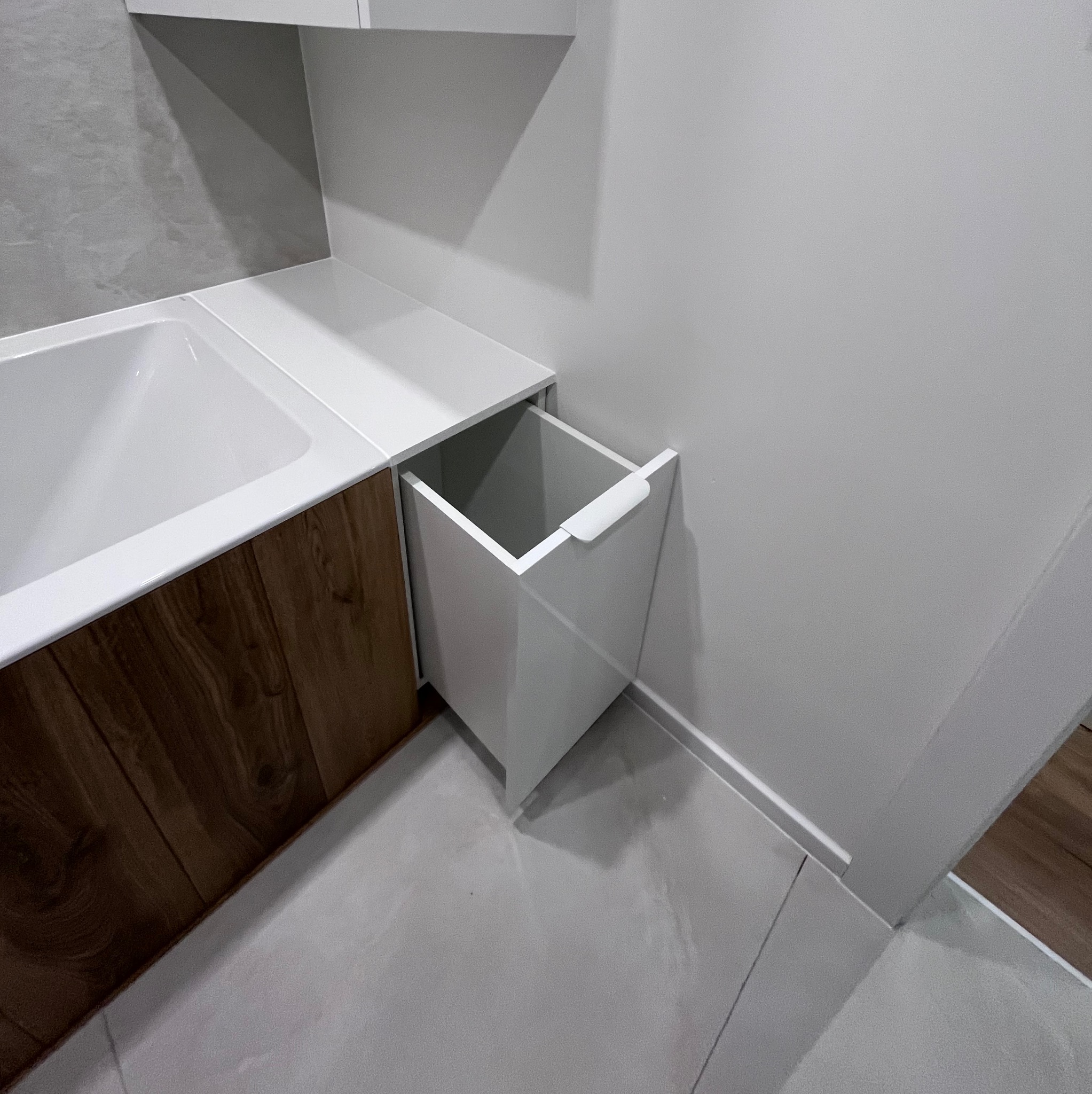 Nowoczesna łazienka z drewnianą zabudową wanny i wysuwaną białą szafką. Minimalistyczny design, szare płytki, jasne wnętrze. Funkcjonalne rozwiązanie.