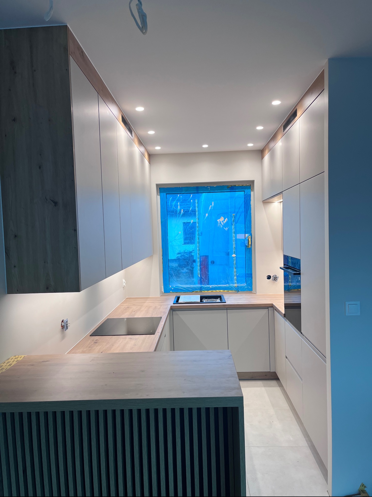 Nowoczesna kuchnia w odcieniach bieli i drewna z wyspą. Okno zabezpieczone folią ochronną. Oświetlenie punktowe podkreśla minimalistyczny design.