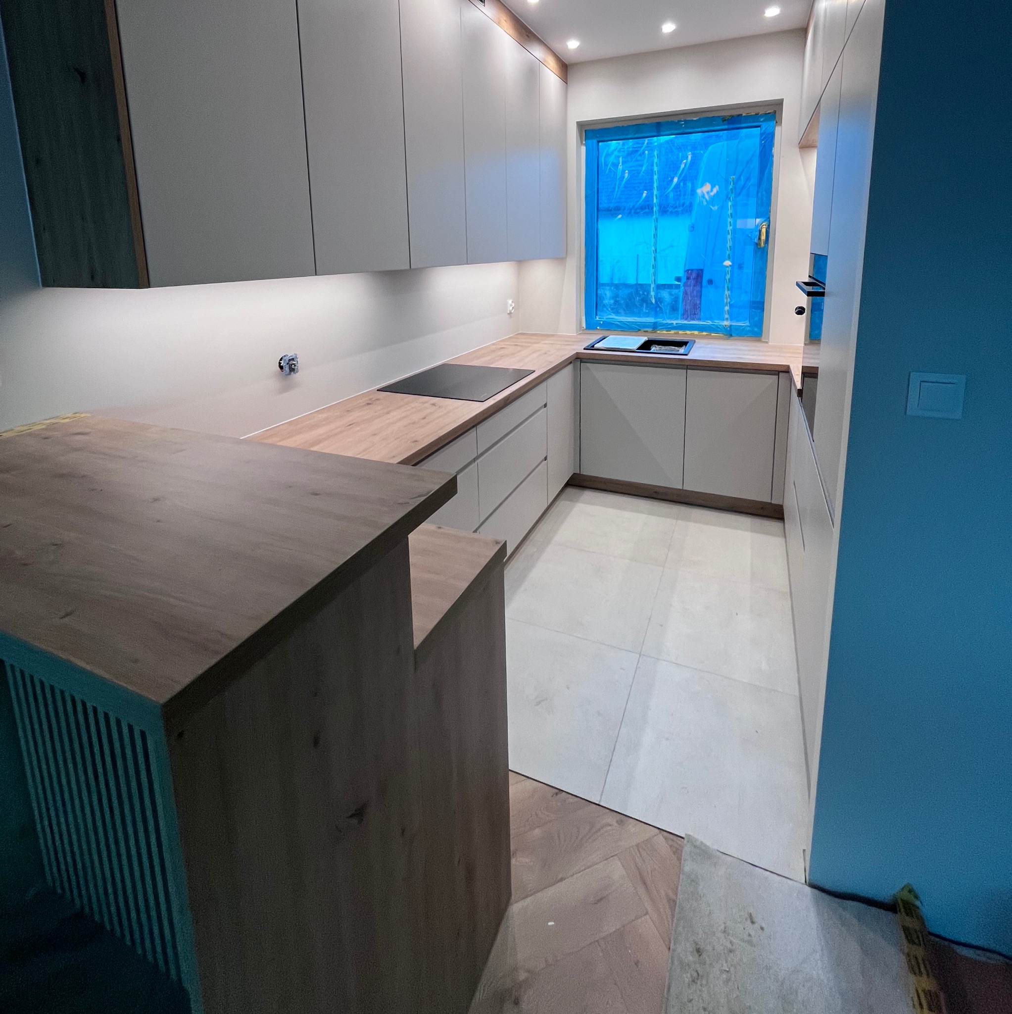 Jasna kuchnia z drewnianym blatem i wyspą. Okno zabezpieczone folią. Nowoczesne oświetlenie podszafkowe. Parkiet ułożony w jodełkę. Minimalistyczny design.