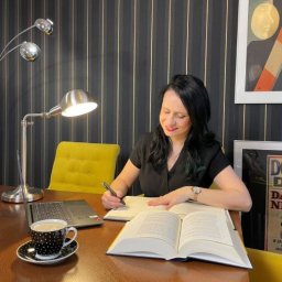 Adwokaci Ireneusz Sachanbiński, Olga Sachanbińska-Dobrzyńska - Kobieta w czarnej bluzce, siedząca za biurkiem z otwartą książką, laptopem, filiżanką kawy w kropki i lampą, pisząca coś w notatniku.