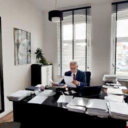 Adwokaci Ireneusz Sachanbiński, Olga Sachanbińska-Dobrzyńska - Wnętrze biura z adwokatem w garniturze, analizującym dokumenty przy czarnym biurku, na którym leżą sterty papierów i laptop. W tle okna z żaluzjami i widokiem na miasto oraz regał z dekoracjami.