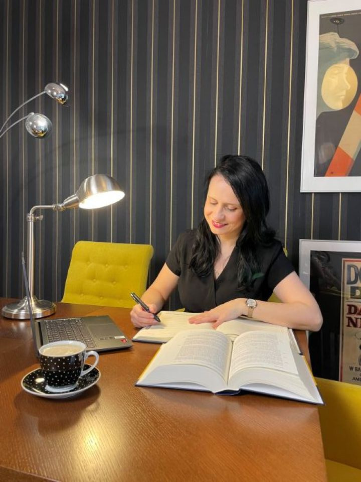 Kobieta w czarnej bluzce, siedząca za biurkiem z otwartą książką, laptopem, filiżanką kawy w kropki i lampą, pisząca coś w notatniku.