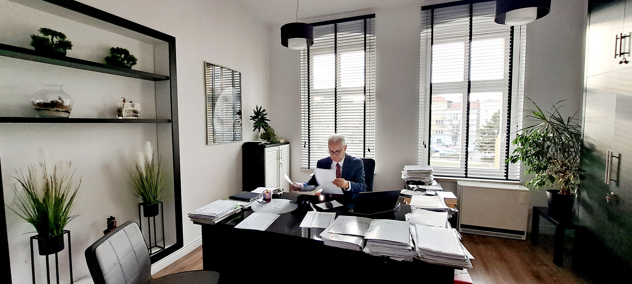 Wnętrze biura z adwokatem w garniturze, analizującym dokumenty przy czarnym biurku, na którym leżą sterty papierów i laptop. W tle okna z żaluzjami i widokiem na miasto oraz regał z dekoracjami.