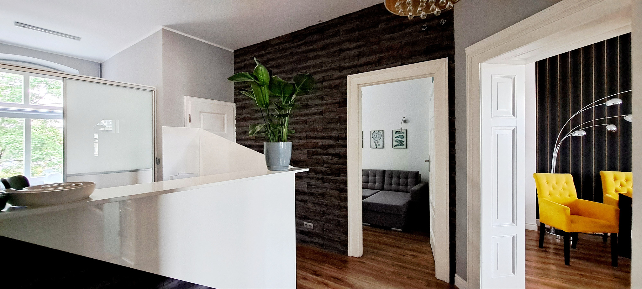 Wnętrze kancelarii: biała recepcja z rośliną doniczkową, fragment ściany z dekoracyjnej cegły, widok na pokój z szarą sofą oraz pokój z żółtymi fotelami i lampą łukową.