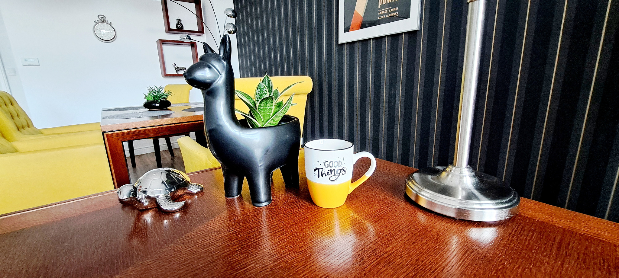 Aranżacja wnętrza z drewnianym stołem, na którym stoi czarna doniczka w kształcie lamy z rośliną, srebrna figurka żółwia i kubek z napisem 'Good Things', w tle żółty fotel i ściana w pionowe pasy.