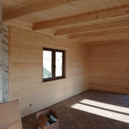 Dawid Ziera - Wnętrze domu w stylu skandynawskim z drewnianymi ścianami i sufitem. Widoczne okno, podłoga z wzorem, kartony i puszki z farbą. Jasne, naturalne oświetlenie.