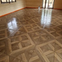 Dawid Ziera - Wnętrze z drewnianą podłogą ułożoną we wzór parkietu, widoczne ściany z drewnianych paneli i okno. Podłoga zabezpieczona pomarańczową taśmą.