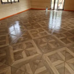 Wnętrze z drewnianą podłogą ułożoną we wzór parkietu, widoczne ściany z drewnianych paneli i okno. Podłoga zabezpieczona pomarańczową taśmą.