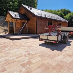 Dawid Ziera - Drewniany dom z ciemnym dachem i brązowymi rynnami, zadaszony taras, kostka brukowa na podwórku, przyczepa z narzędziami. Słoneczny dzień, zielone drzewa w tle.