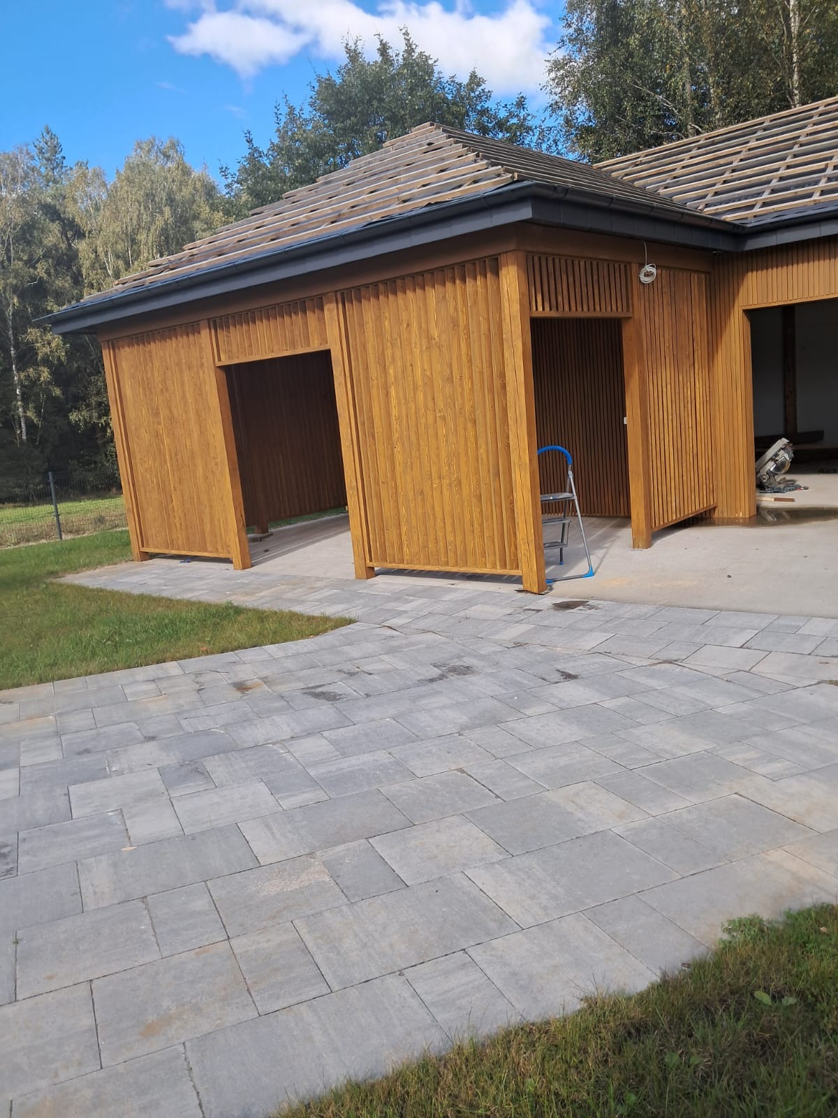 Drewniana konstrukcja z otwartymi wjazdami, w trakcie budowy dachu, z niebieską drabinką wewnątrz, na tle kostki brukowej i zieleni. Słoneczny dzień.