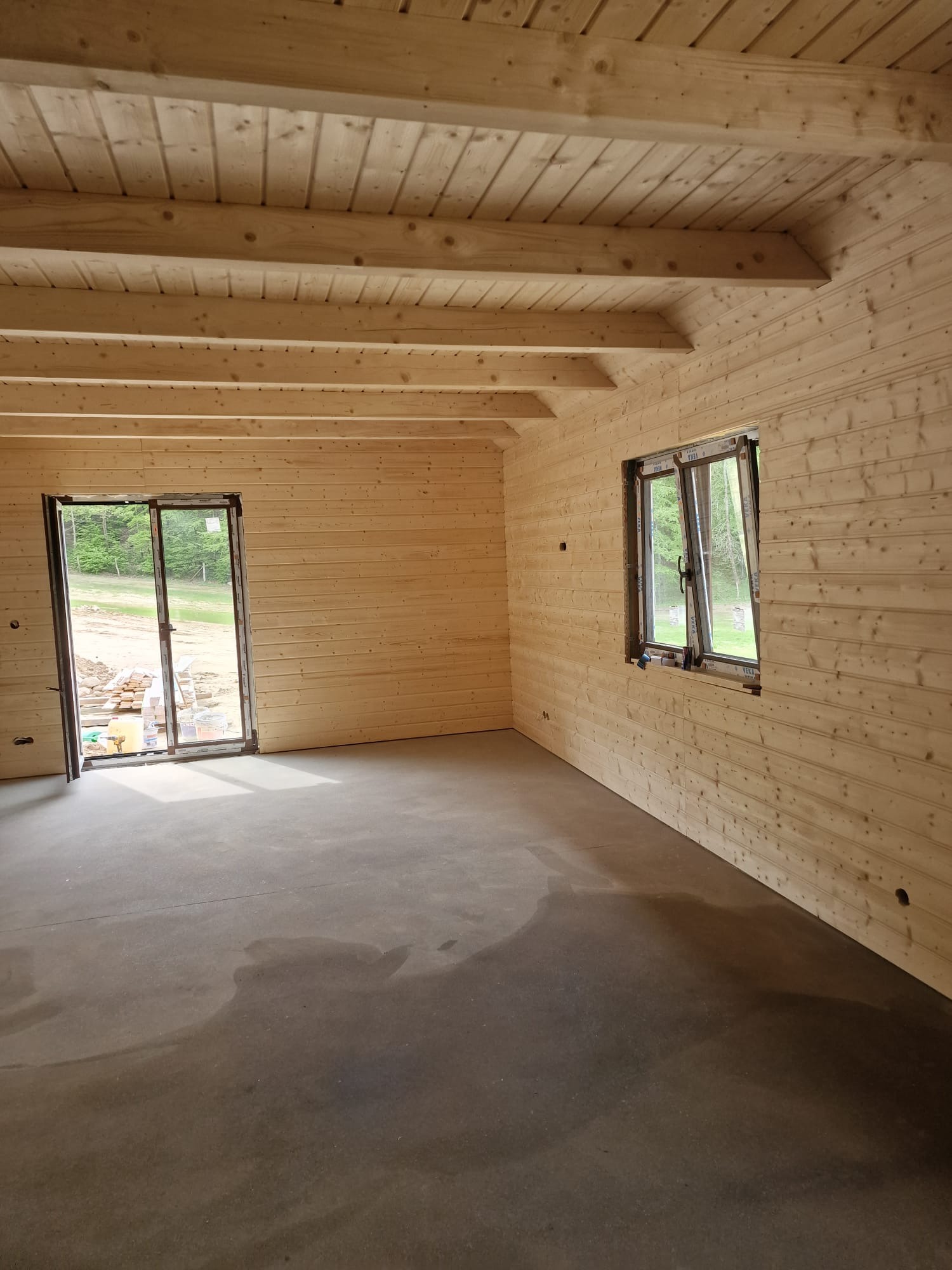 Wnętrze domu w trakcie budowy z drewnianymi ścianami, sufitem z belek i betonową podłogą. Widoczne okno i drzwi balkonowe z widokiem na zieleń.