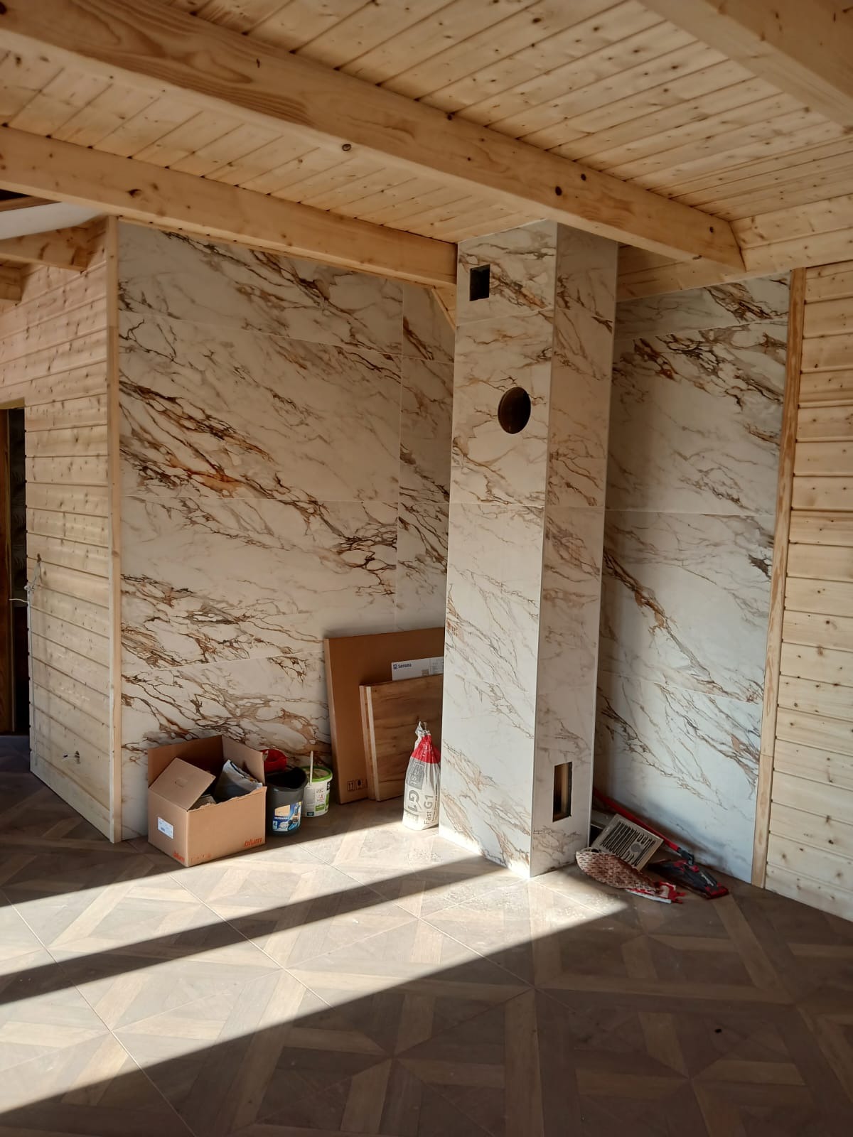 Wnętrze w trakcie wykończenia: drewniane ściany, marmurowe płytki na ścianach i kominie, wzorzysta podłoga. Widoczne narzędzia i materiały budowlane.