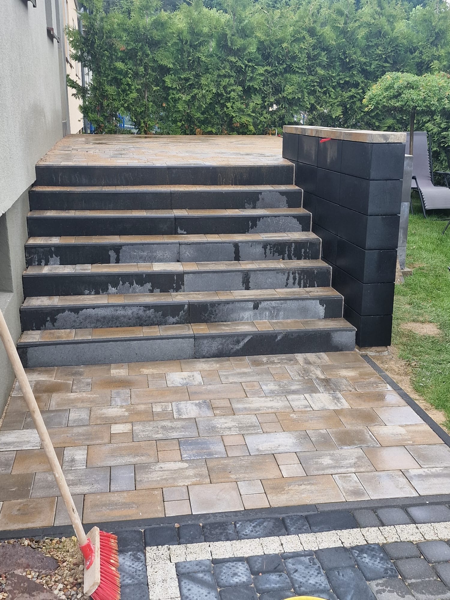 Ułożone schody z kostki brukowej w odcieniach szarości i beżu, prowadzące do domu. Widoczny fragment chodnika z kostki oraz zieleni w tle.