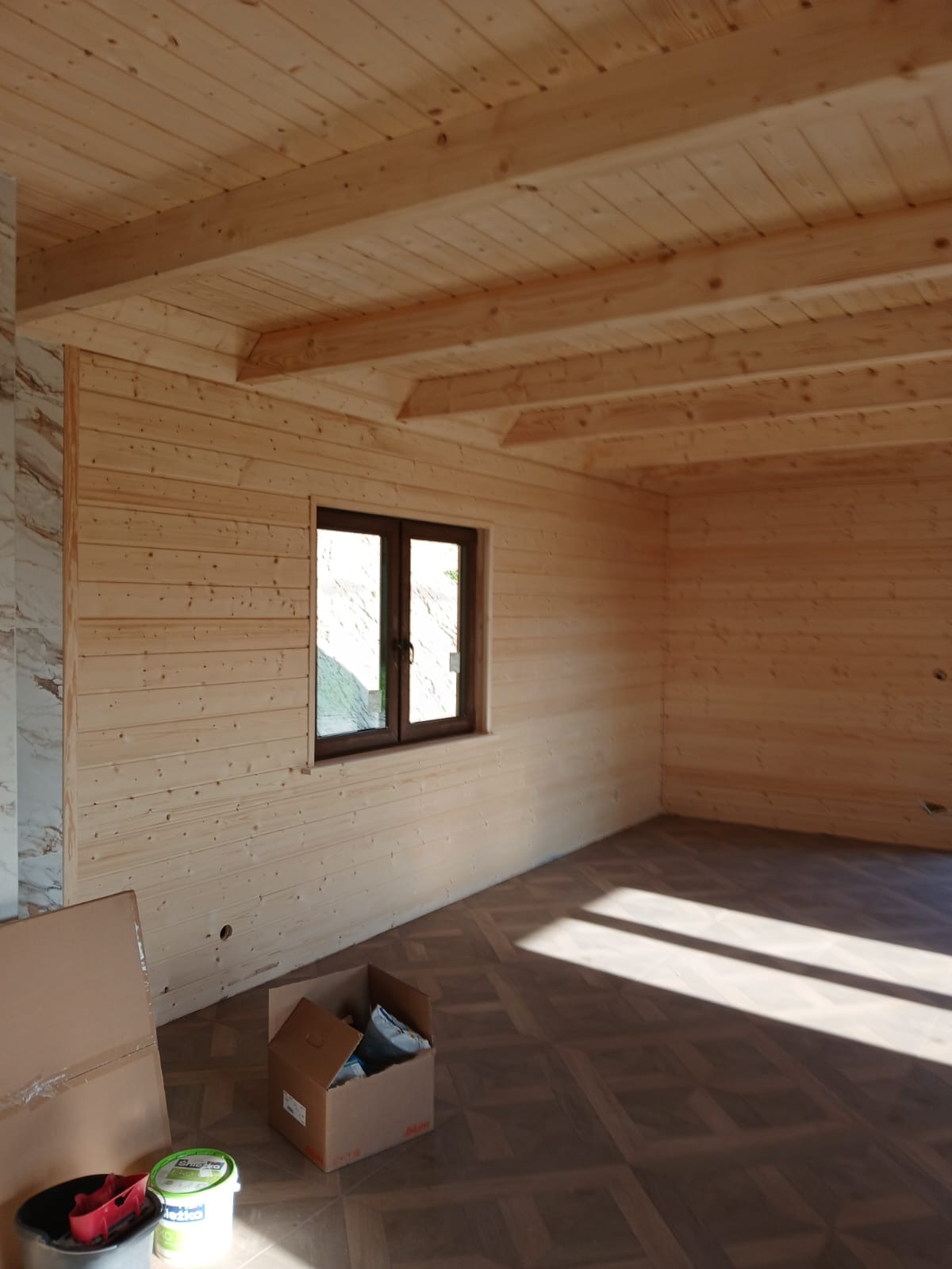 Wnętrze domu w stylu skandynawskim z drewnianymi ścianami i sufitem. Widoczne okno, podłoga z wzorem, kartony i puszki z farbą. Jasne, naturalne oświetlenie.