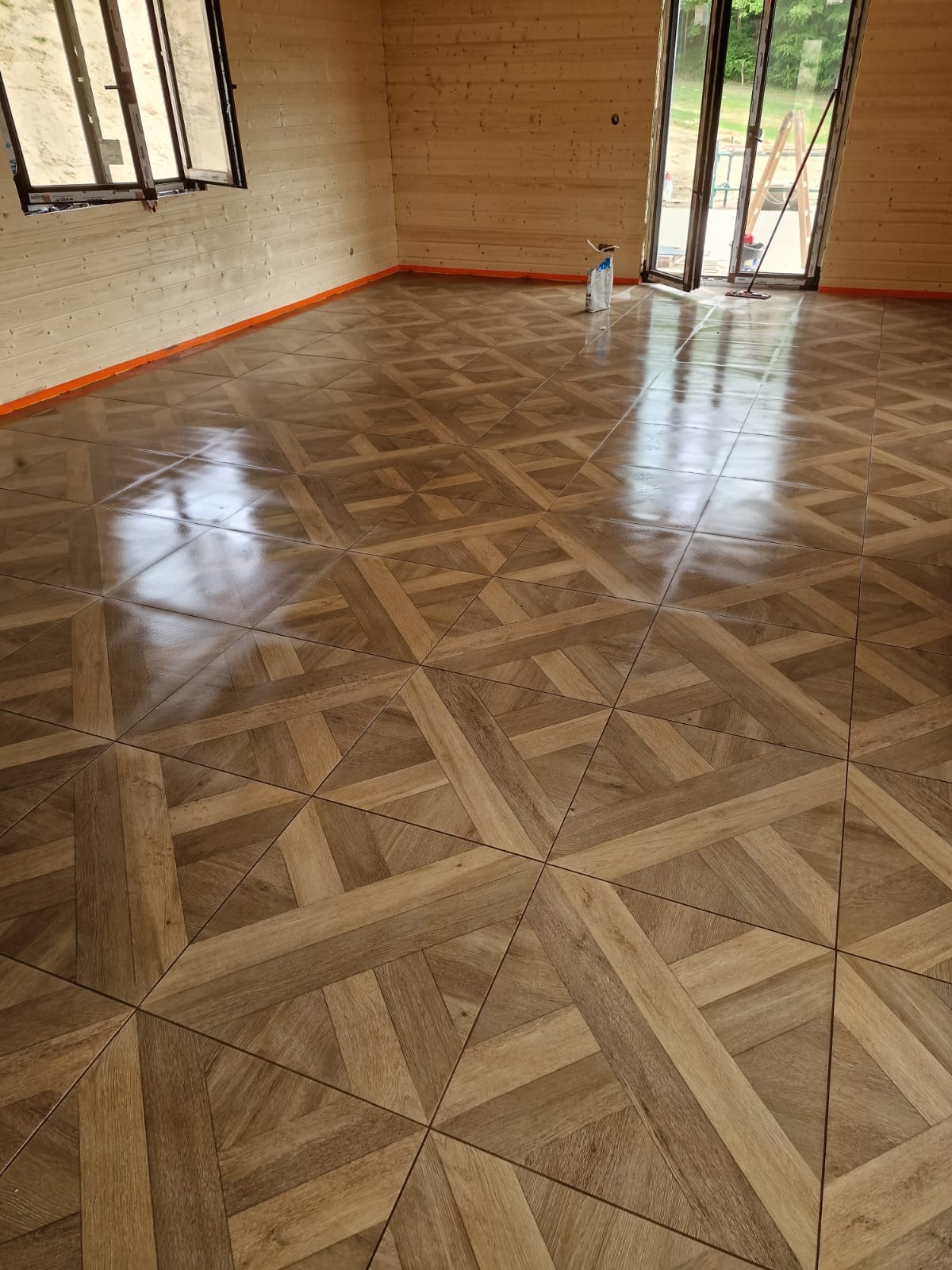 Wnętrze z drewnianą podłogą ułożoną we wzór parkietu, widoczne ściany z drewnianych paneli i okno. Podłoga zabezpieczona pomarańczową taśmą.