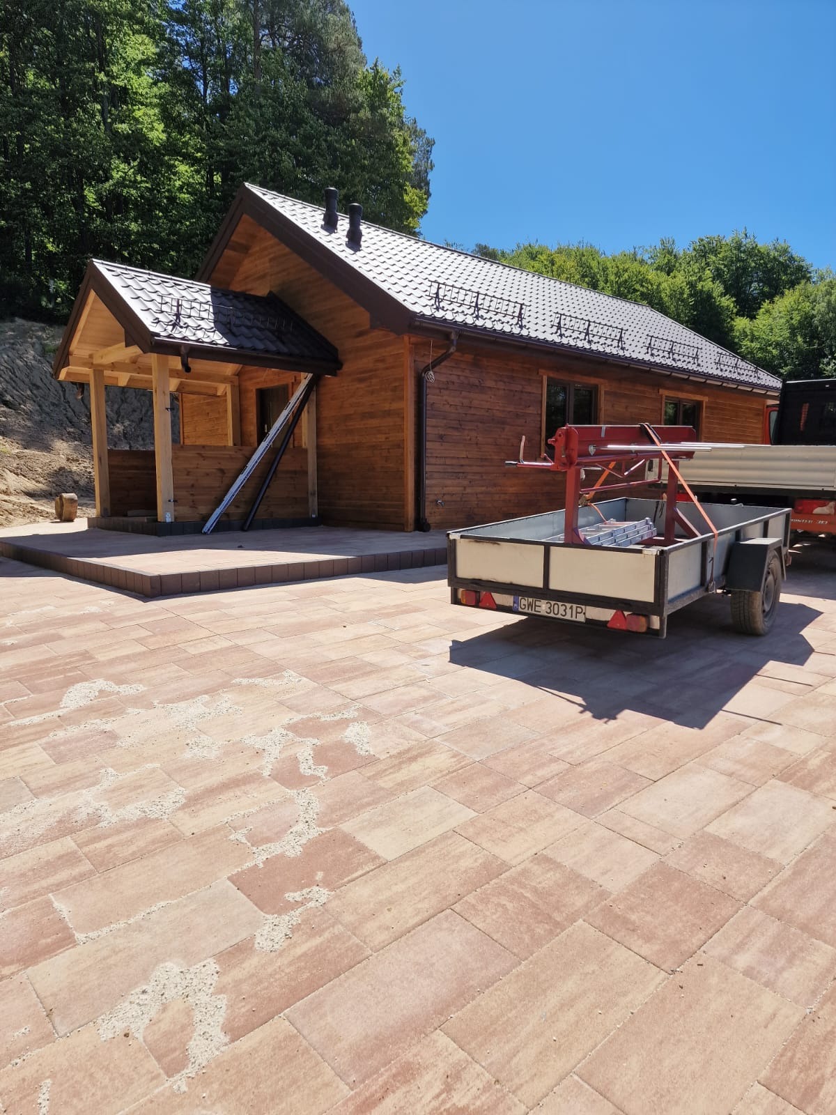 Drewniany dom z ciemnym dachem i brązowymi rynnami, zadaszony taras, kostka brukowa na podwórku, przyczepa z narzędziami. Słoneczny dzień, zielone drzewa w tle.