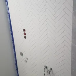 Ściana łazienki wyłożona białymi, błyszczącymi płytkami ułożonymi w jodełkę, widoczne otwory na armaturę, dwa wiadra i akcesoria glazurnicze, częściowo widoczna kabina prysznicowa z niebieską taśmą...