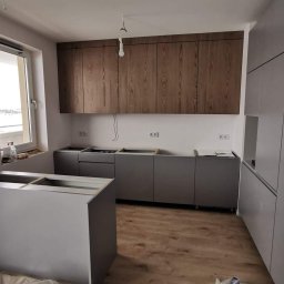 Usługi wykończeniowe i naprawcze - Wnętrze kuchni w trakcie montażu: szare szafki dolne i wysokie zabudowy, drewniane szafki wiszące, widoczne przyłącza elektryczne i fragment okna z widokiem na zewnątrz.