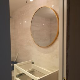 Usługi wykończeniowe i naprawcze - Niedokończona łazienka z beżowymi płytkami, okrągłym lustrem w złotej ramie i niezmontowaną szafką pod umywalkę; widoczne niedokończone wykończenie ościeżnicy i prowizoryczne oświetlenie.