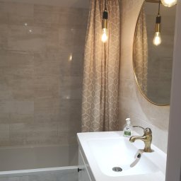 Usługi wykończeniowe i naprawcze - Wykończona łazienka w neutralnych kolorach z beżową zasłoną prysznicową w geometryczny wzór, złotą armaturą i okrągłym lustrem ze złotą ramą.