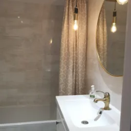 Wykończona łazienka w neutralnych kolorach z beżową zasłoną prysznicową w geometryczny wzór, złotą armaturą i okrągłym lustrem ze złotą ramą.