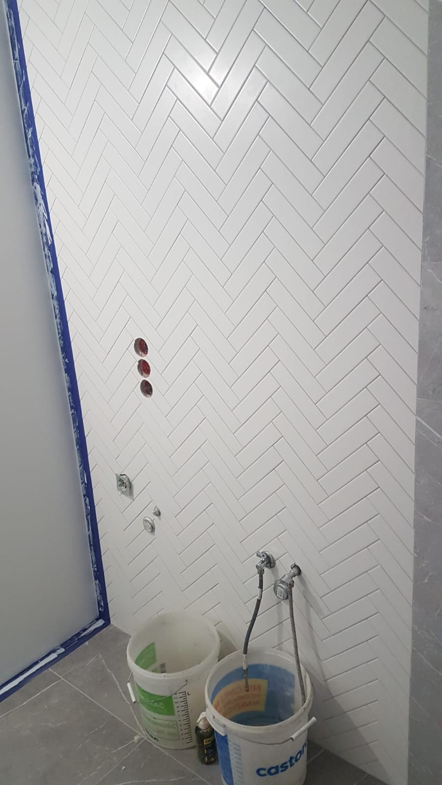 Ściana łazienki wyłożona białymi, błyszczącymi płytkami ułożonymi w jodełkę, widoczne otwory na armaturę, dwa wiadra i akcesoria glazurnicze, częściowo widoczna kabina prysznicowa z niebieską taśmą...