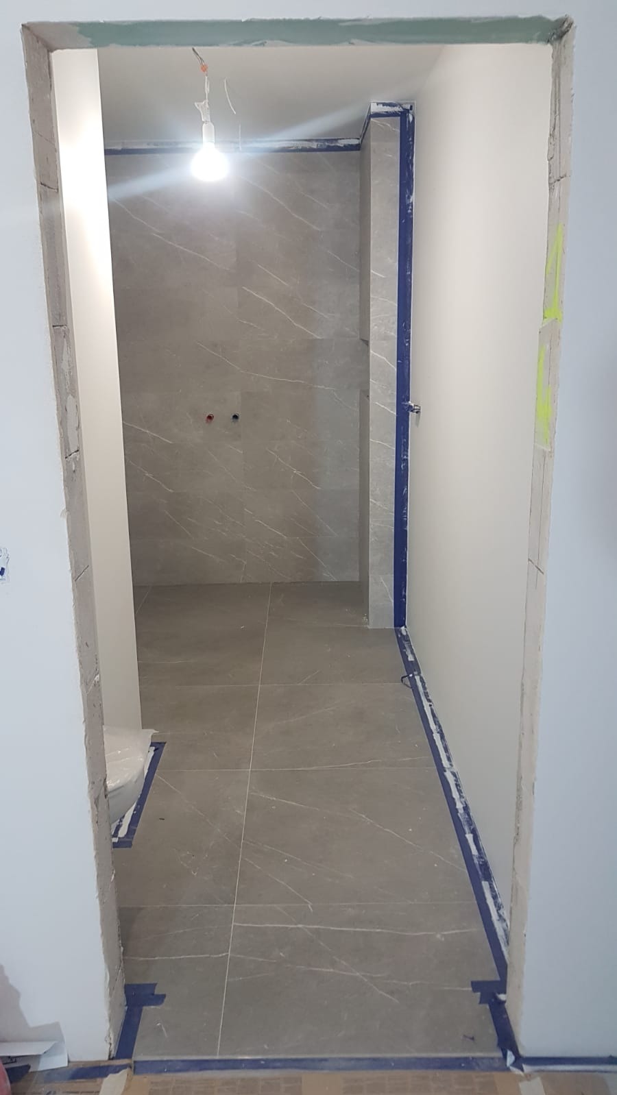 Wnętrze łazienki w trakcie remontu: szare płytki imitujące marmur na podłodze i ścianach, zabezpieczone niebieską taśmą malarską, surowy otwór drzwiowy.