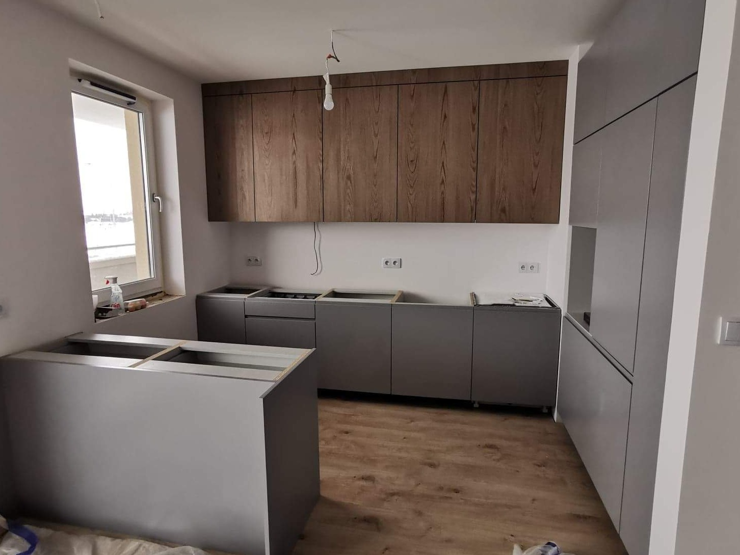 Wnętrze kuchni w trakcie montażu: szare szafki dolne i wysokie zabudowy, drewniane szafki wiszące, widoczne przyłącza elektryczne i fragment okna z widokiem na zewnątrz.